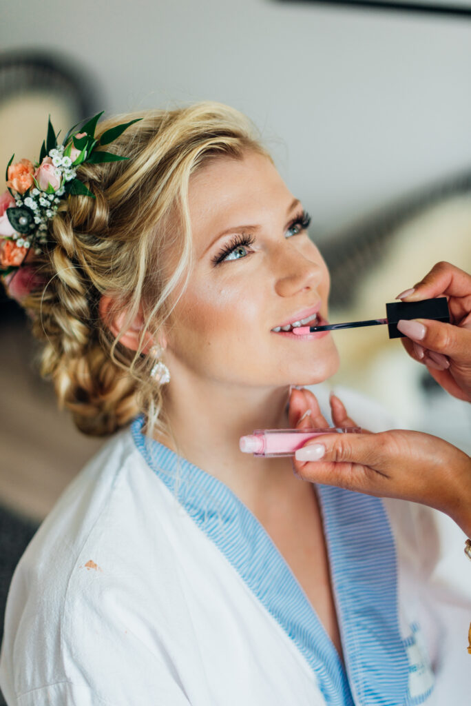 Elin Hang sminkar en blivande brud. Applicerar läppglas. Bruden sitter på med ett leende på läpparna och blommor i håret med med en inbakad fläta.